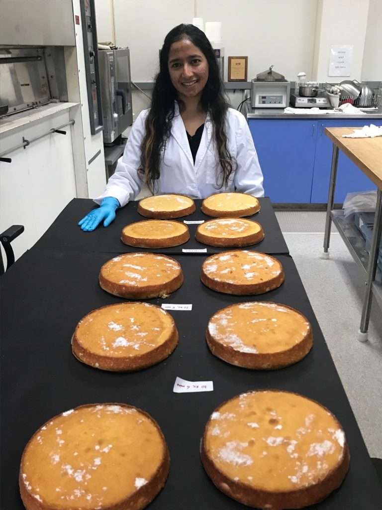 A woman in a lab coat and blue glove smiles while standing behind a table with eight round sponge cakes, each labeled with a small tag, in a laboratory setting.
