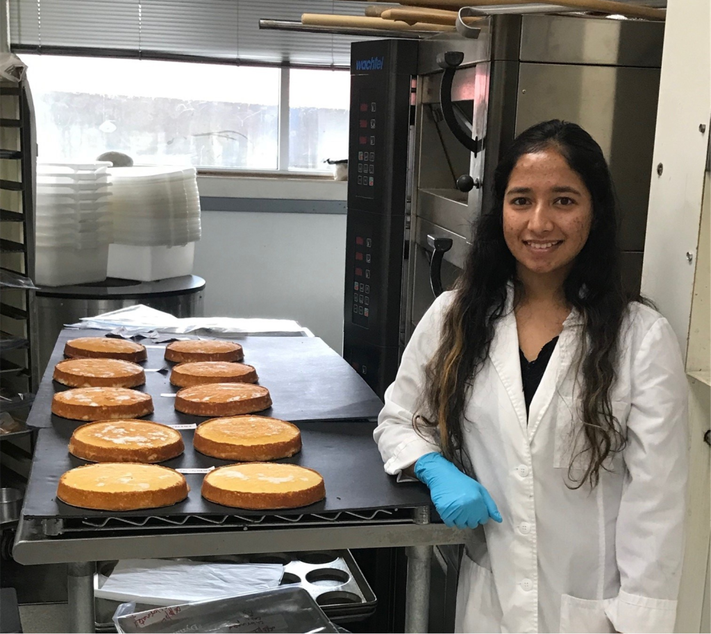A woman in a white lab coat and blue glove stands beside trays of round cakes on a rack in a kitchen or bakery, smiling at the camera. Baking equipment and stacked containers are visible in the background.