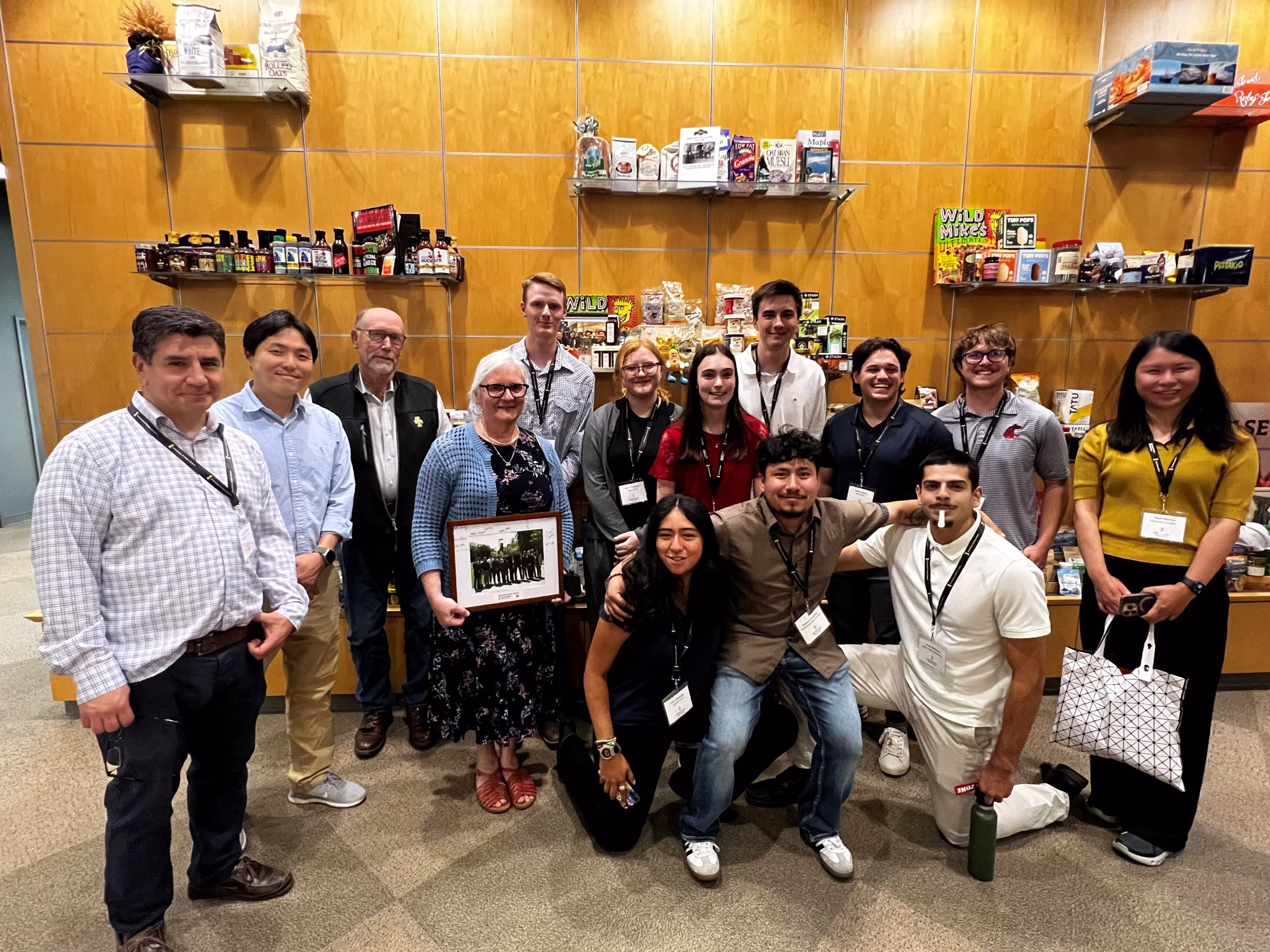 A group of fourteen people pose and smile for a photo in front of shelves filled with snacks and products. One person in the front row holds a framed photo. Most are wearing conference badges.