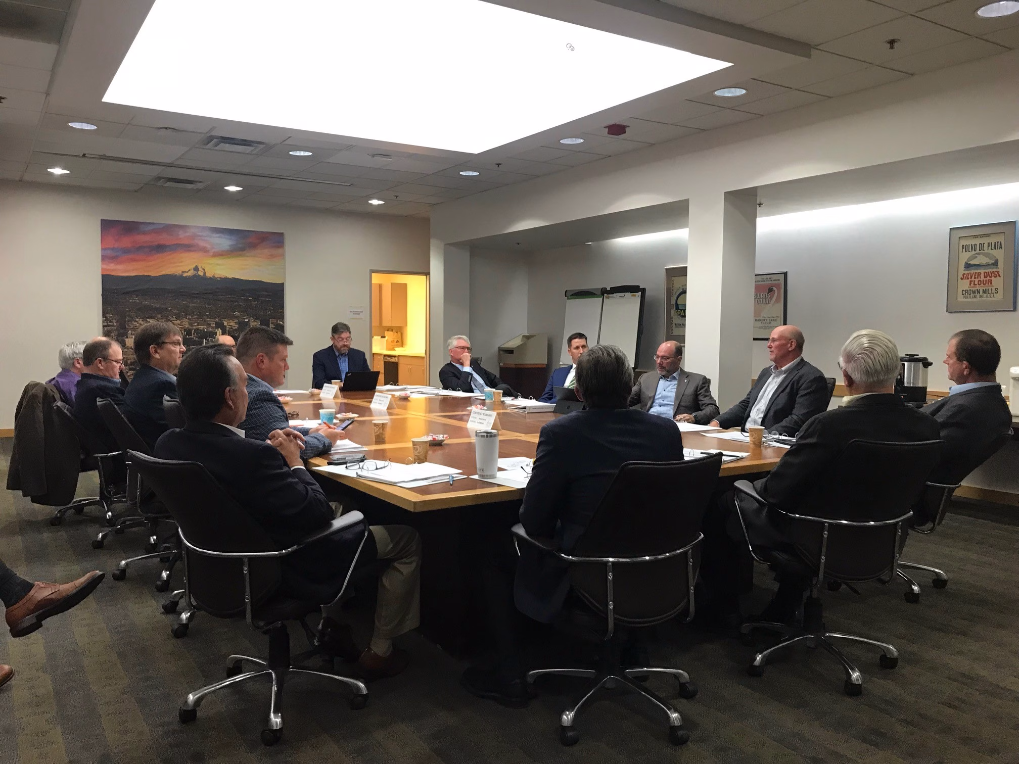 A group of men in business attire sit around a large conference table in a well-lit meeting room, engaged in discussion. Papers and coffee cups are on the table, and office artwork hangs on the walls.