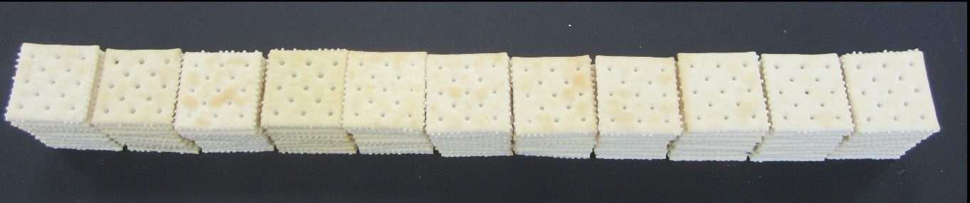 Stacks of square saltine crackers arranged in a neat horizontal row on a dark, flat surface. Each stack consists of several crackers placed directly on top of one another.