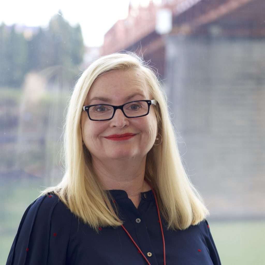A woman with long blonde hair and glasses smiles at the camera. She is wearing a dark blouse and a red lanyard, with a blurred outdoor scene and a bridge in the background.