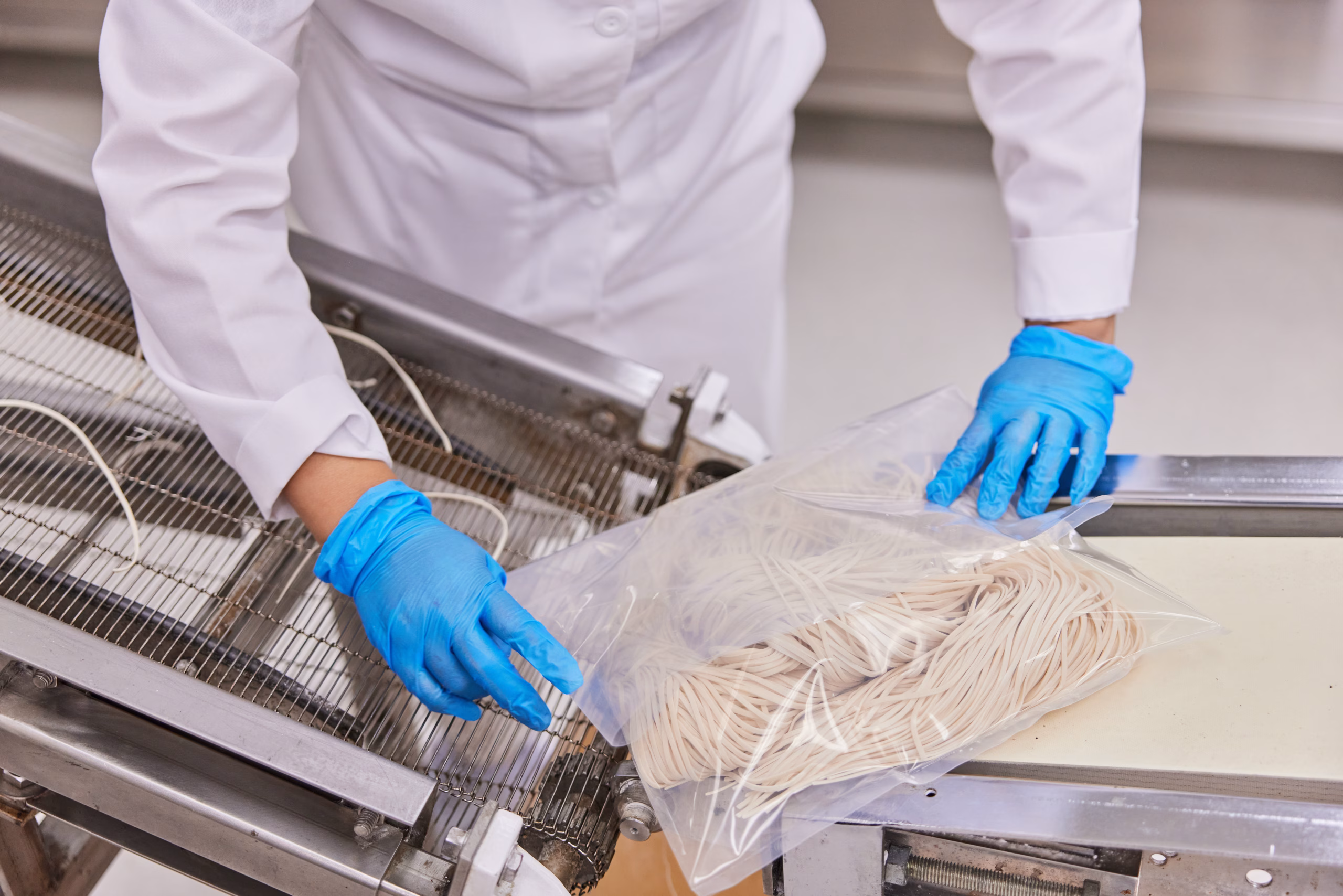 A person wearing a white lab coat and blue gloves places uncooked noodles into a clear plastic bag on a food production assembly line.