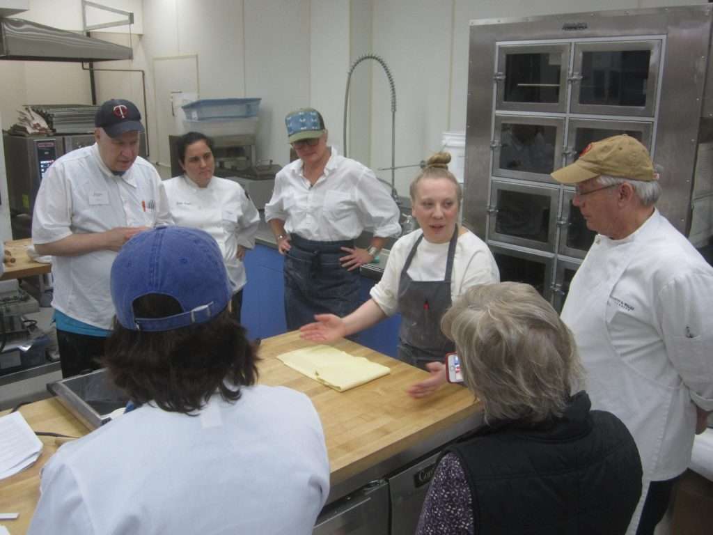 A group of people in chef uniforms gather around a kitchen island as a woman demonstrates a technique with dough, engaging the group in a commercial kitchen setting.