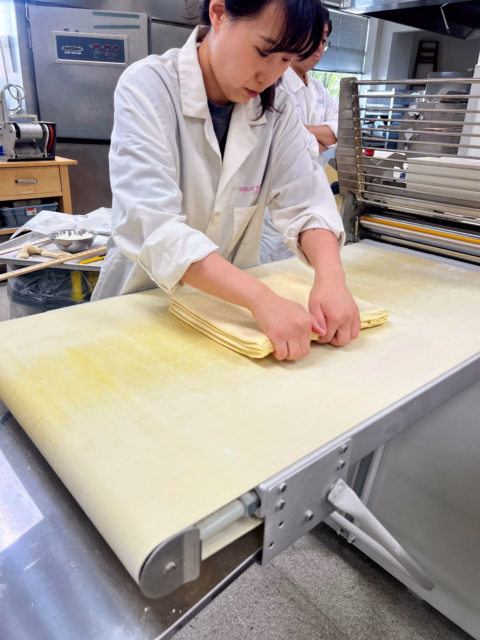 A person in a white lab coat folds dough sheets on a large worktable in a professional kitchen, preparing it for processing with a dough sheeter machine.