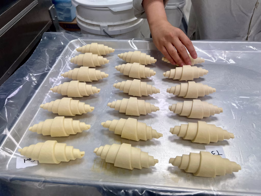 A person places uncooked croissants on a baking tray lined with plastic, preparing them for baking. Multiple rows of neatly arranged dough are visible.