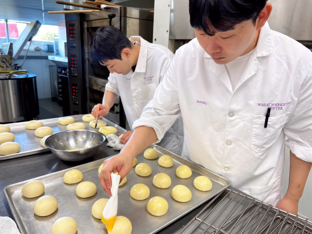 Two people in white uniforms brush egg wash onto dough balls arranged on baking trays in a commercial kitchen. One focuses on the tray in front, while the other works on a tray in the background.