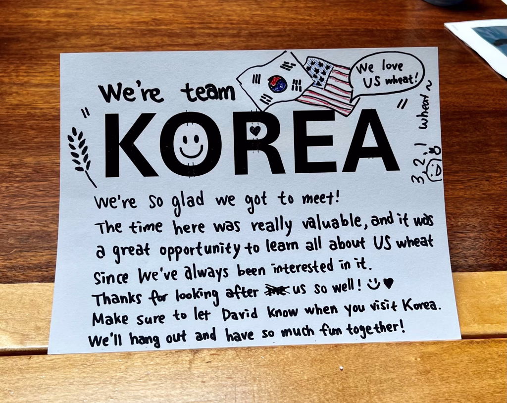 A handwritten note on a table reads: Were team KOREA :) We love US wheat! Were so glad we got to meet! The time here was valuable... Thanks for looking after us! Well hang out and have fun together! With doodles of flags and hearts.