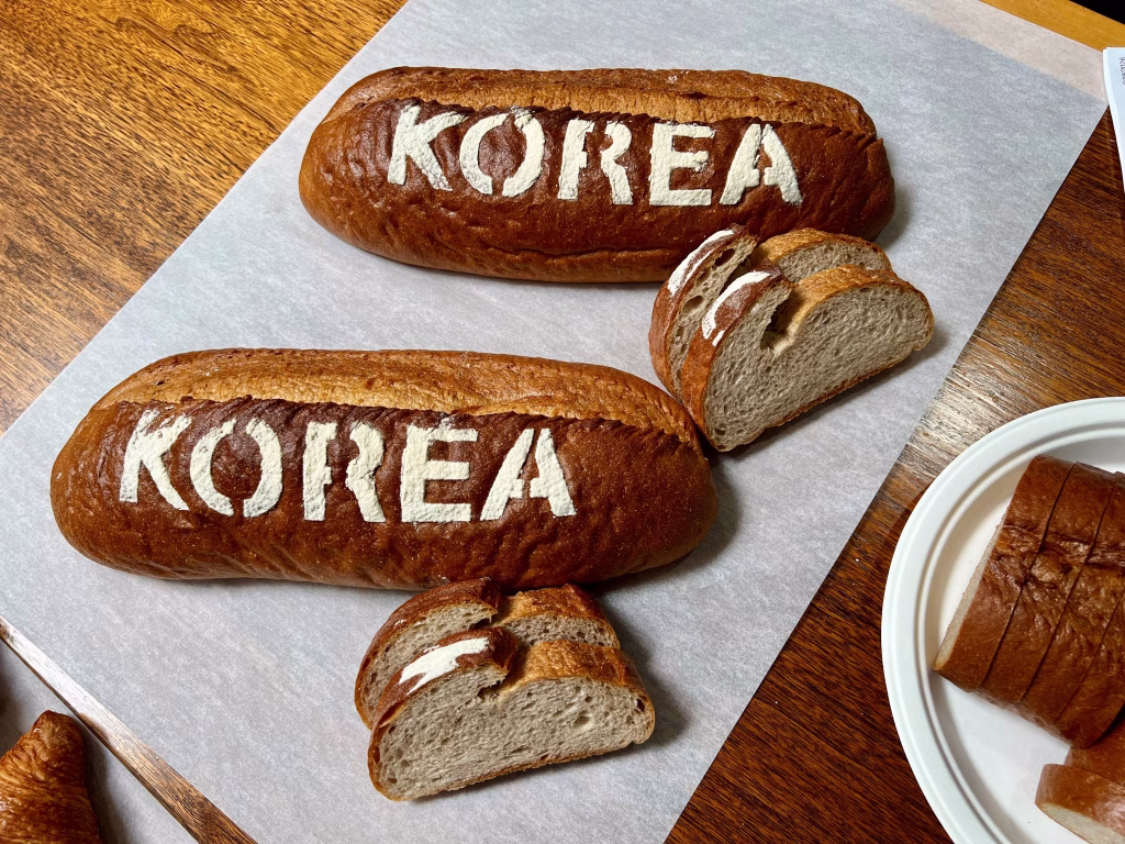 Two loaves of brown bread with the word KOREA stenciled on top in white flour, one partially sliced, displayed on parchment paper on a wooden surface. Slices of bread are placed nearby.
