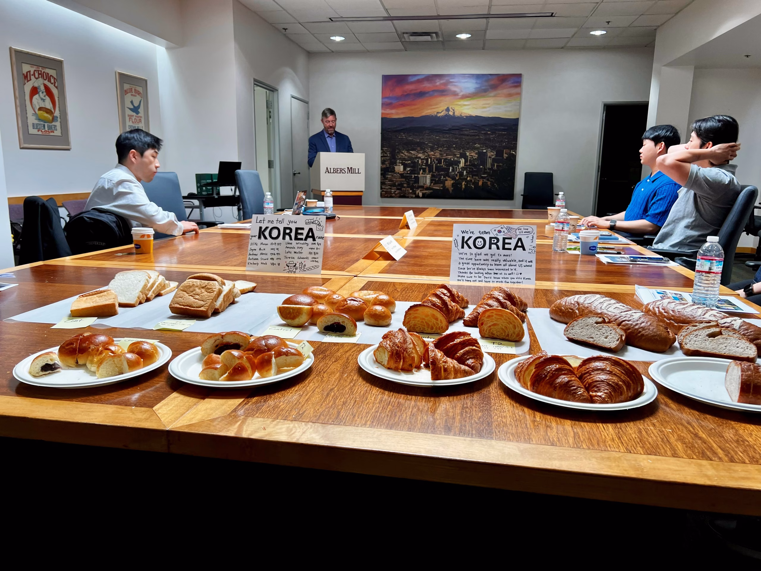 A conference room with people seated around a table, assorted breads and pastries labeled Korea in the foreground, and a man speaking at a podium in the background. A large cityscape photo hangs on the wall.