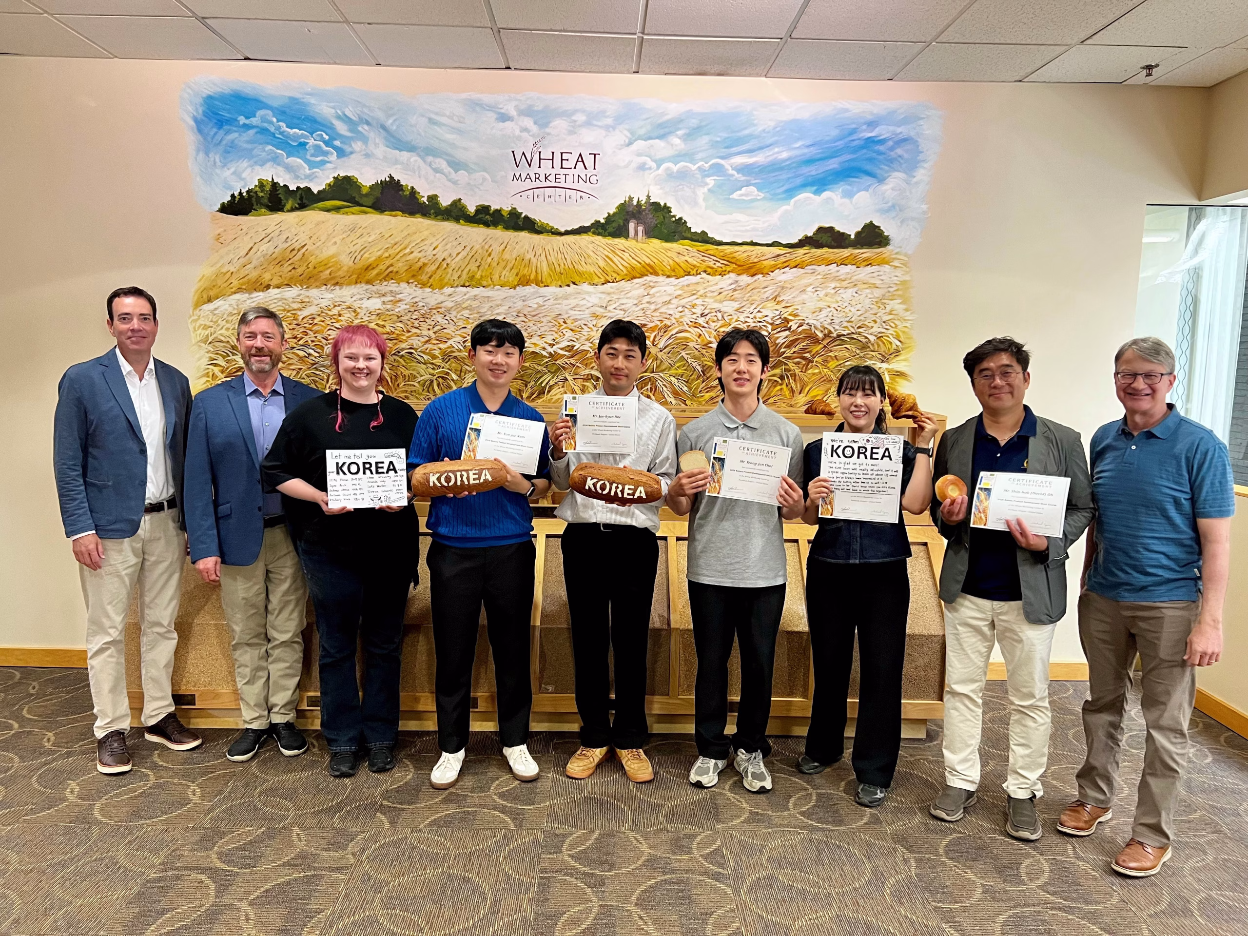 A group of nine people stands indoors in front of a mural of a wheat field. Four students in the center hold certificates and bread labeled Korea, flanked by five adults, all smiling at the camera.