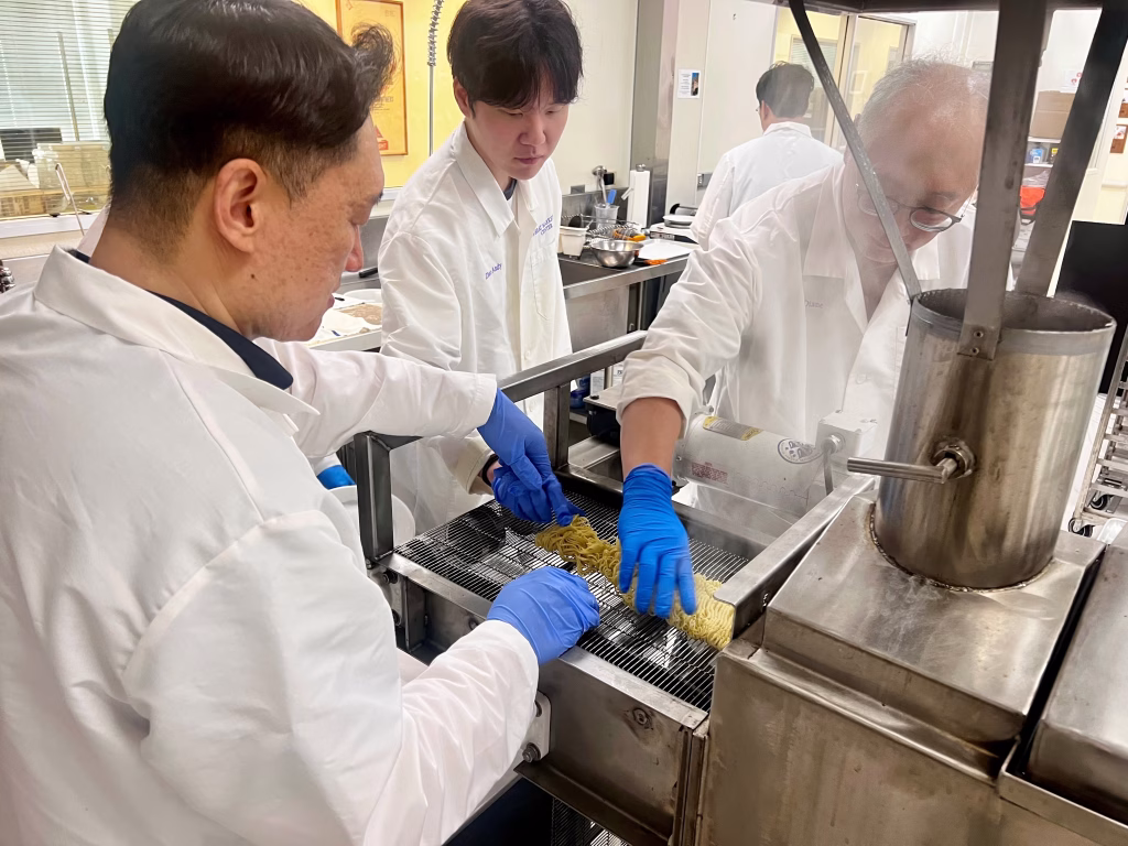 Three people in lab coats and blue gloves work together, placing noodles onto a metal conveyor near industrial kitchen equipment in a food processing laboratory.