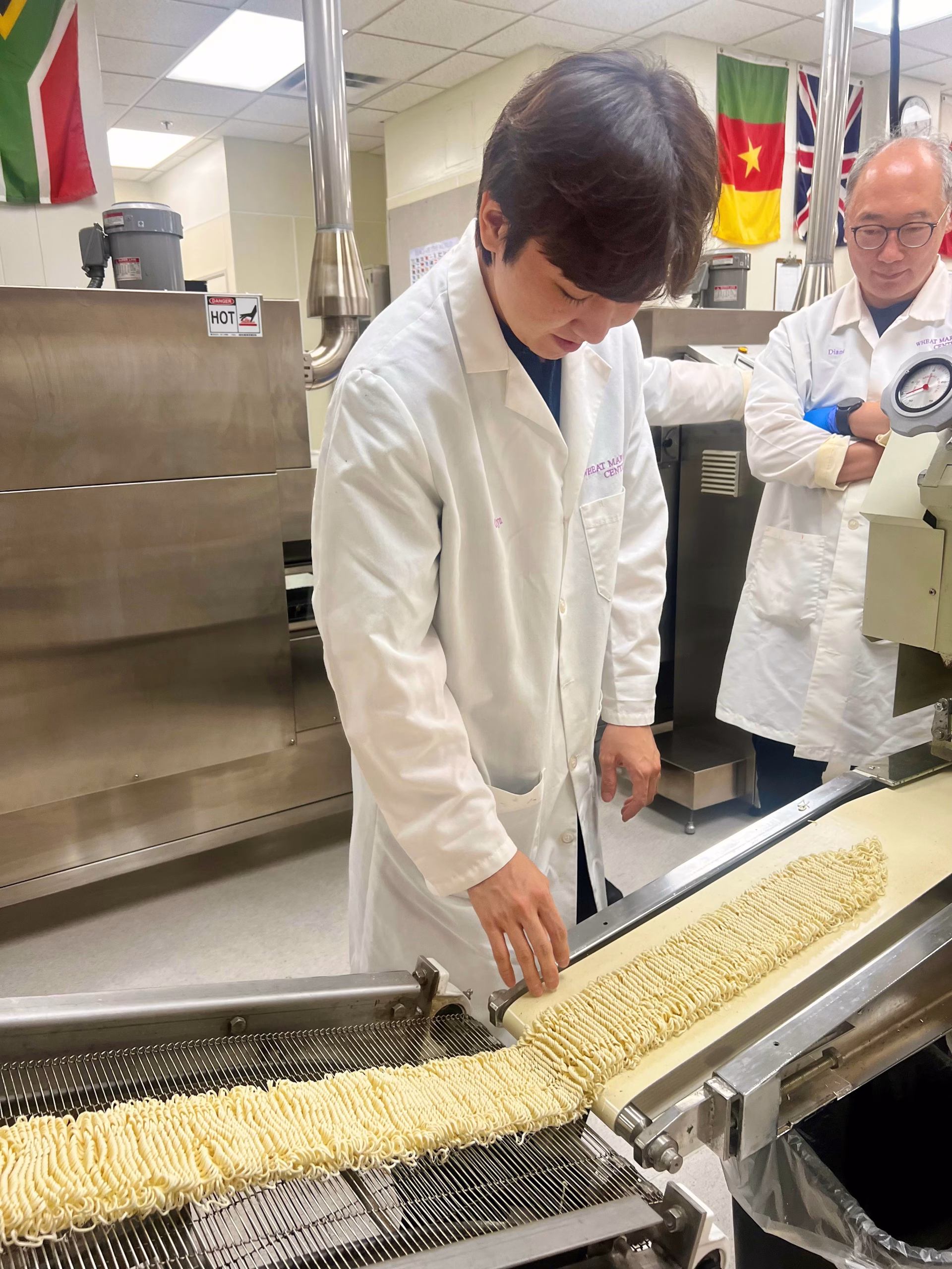 Two people in lab coats observe as one adjusts a long strip of uncooked ramen noodles on a conveyor in a food processing lab, with international flags hanging in the background.