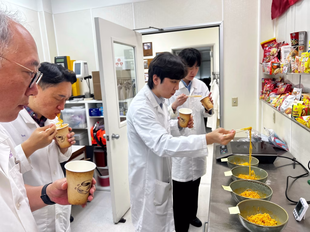 Four people in white lab coats taste and examine bowls of instant noodles in a lab. Shelves with snack packages and equipment are visible. One person lifts noodles with chopsticks, while others eat or observe.