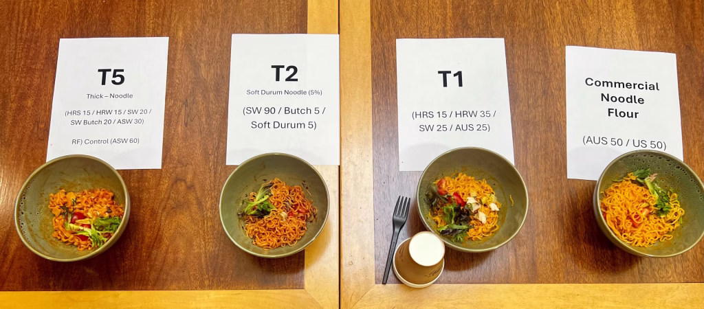 Three bowls of instant noodles labeled T5, T2, and T1, each placed on paper with product info. T1 includes a cup of white liquid. Noodles have various toppings and seasonings, and all are on a wooden surface.