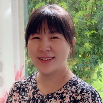 A woman with straight dark hair and bangs smiles indoors, standing near a window with green trees visible outside. She is wearing a floral-patterned top.