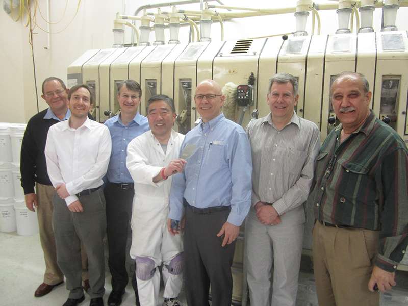 Seven men stand in a row and smile in front of industrial equipment inside a facility; one man is wearing a lab coat, while the others are in business or casual attire.
