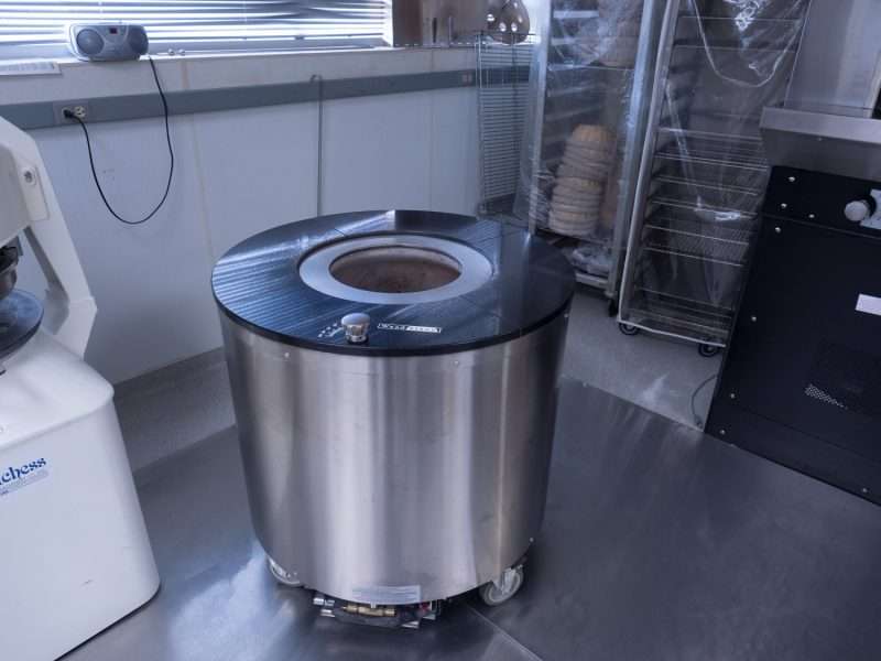 A stainless steel tandoor oven with a black top sits in a commercial kitchen, surrounded by metal surfaces and trays of stacked flatbreads in the background.