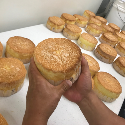 A person holds a round, golden-brown sponge cake with more identical cakes arranged on a white table in the background.